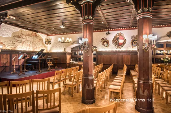 Sala da concerto con sedie in legno, pianoforte a coda, luci da palco e stemmi medievali
